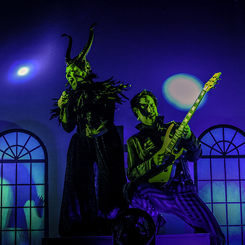 Amid blue and green stage lighting, a costumed female singer screams into a microphone as an electric guitarist kneels beside her.