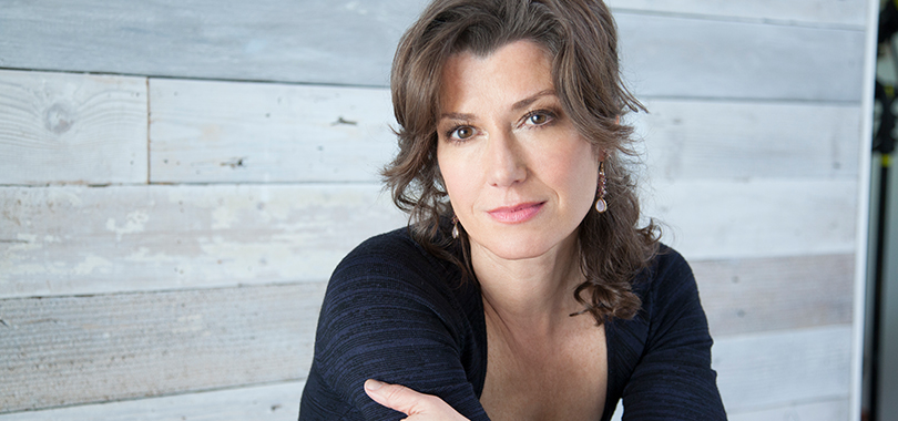 Amy Grant wears a black sweater and poses casually against a wood-panelled wall.