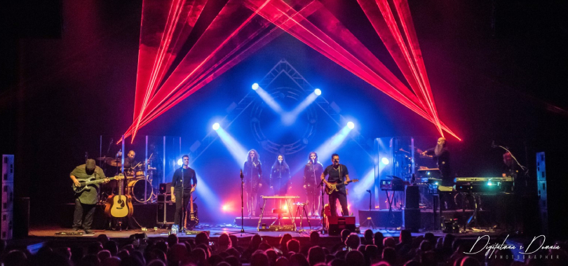 A rock band performs against a stage set featuring triangle-shaped trussing with lighting and red laser lights.