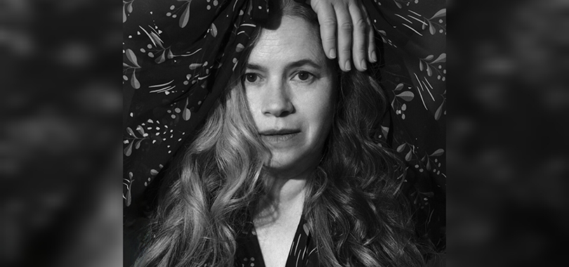Natalie Merchant wears a loose, floral blouse and poses with her hands draped over her head.