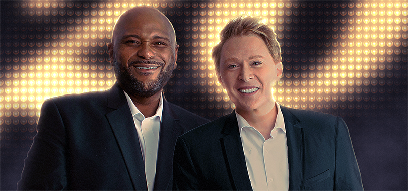 Ruben Studdard and Clay Aiken pose together in dark suits in front of a wall of lights forming the number 20.