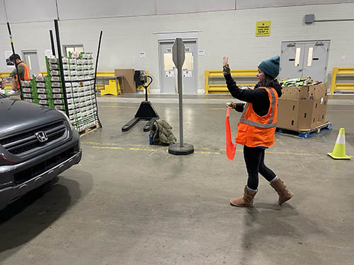 A young woman in an orange safety vest directs traffic inside a Gleaners warehouse.