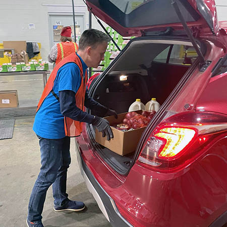 A man in a blue T-shirt and orange safety vest loads food into the back of a car.