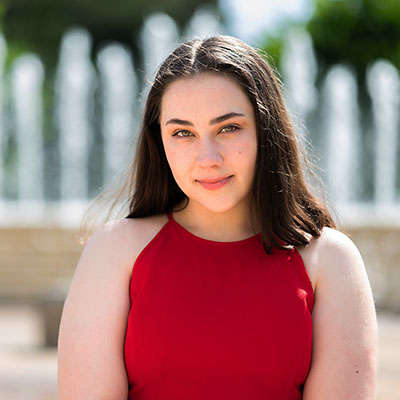 A young woman in a red dress poses before a fountain.