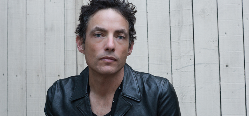 The Wallflowers Jakob Dylan of The Wallflowers wears a black t-shirt under a leather jacket and poses against a plain wood background.