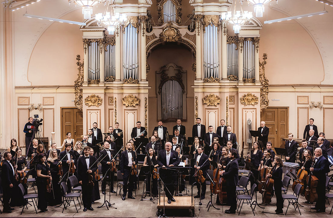 Dressed in black formalwear, the members of the Lviv National Philharmonic Orchestra of Ukraine pose with their instruments on an opulent stage.