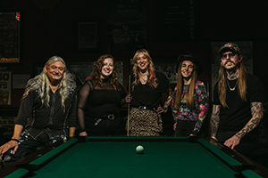 Three women and two men pose for a photo behind a pool table.