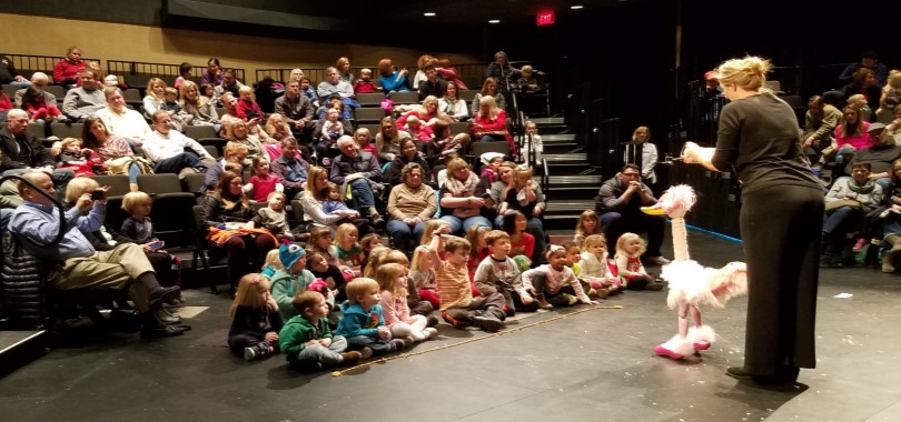 A femalee puppeteer performs with a duck marionette before an audience of children and adults in a black box theater.