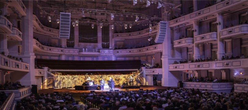 Wide view of a band onstage with a full house at the Palladium concert hall, which is bathed in purple light.