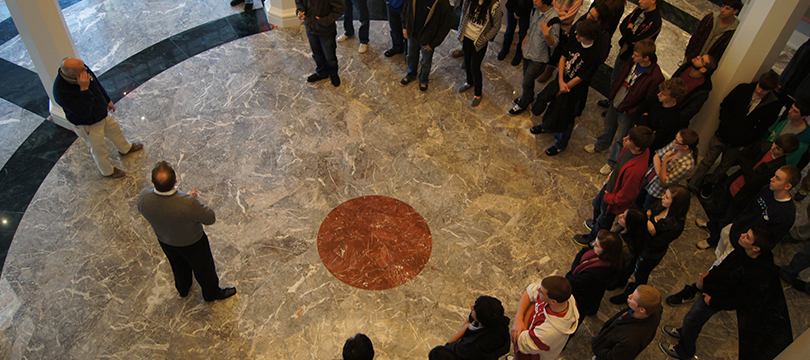 A group of adults gather in a semi-circle around a tour guide on the marble floor of the Palladium's West lobby.
