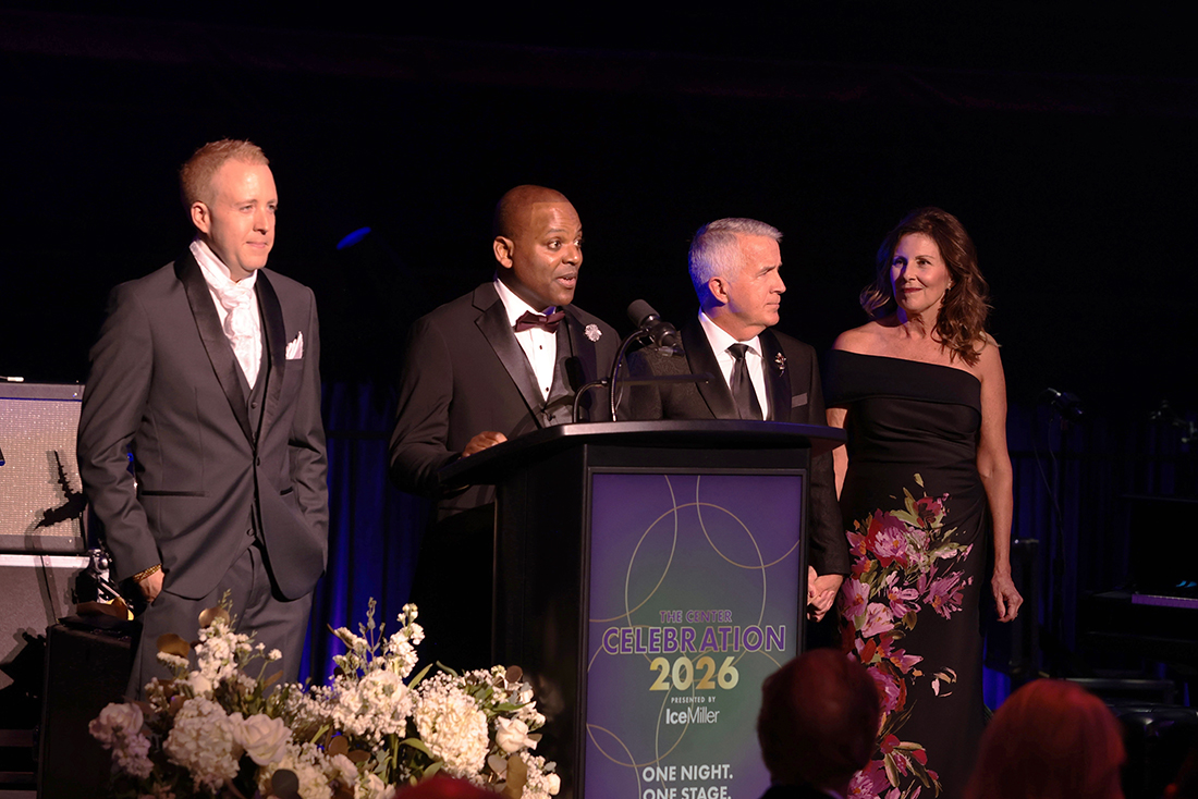 Three men and one woman in formal wear stand around a lectern on a stage.
