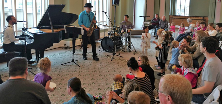Three band members - one on piano, one on tenor saxophone, and one on drumset - perform for a room of children and their parents gathered around on the floor.