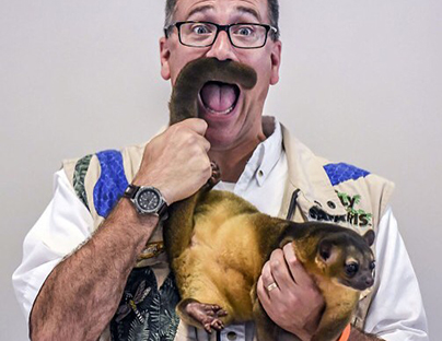A man in glasses holds an animal's tail like a mustache