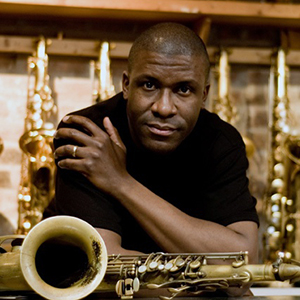 A man in a black shirt poses for portrait leaning against a counter surrounded by saxophones.