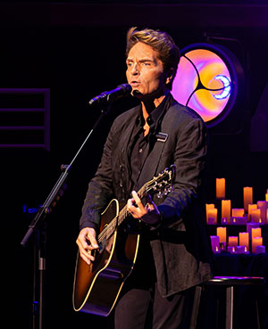 A man in a dark suit strums an acoustic guitar on a stage lit with candles.