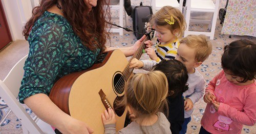 Children gather around a woman with an acoustic guitar.