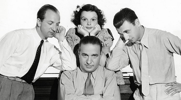 Composer Walter Jurmann, actress-singer Judy Garland and composer Bronislaw Kaper pose around songwriter Gus Kahn in a black-and-white publicity photo..
