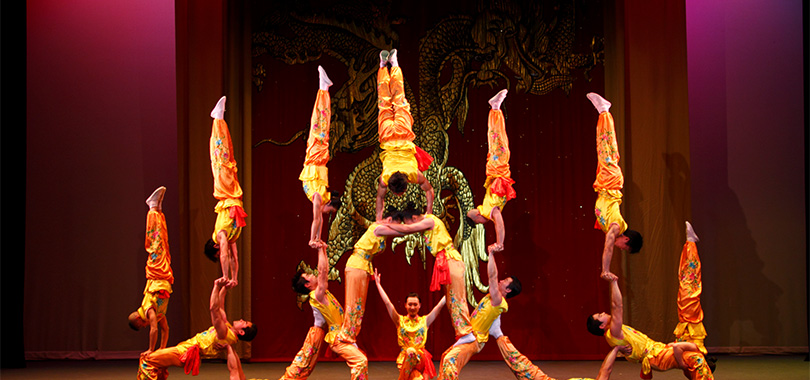 Fifteen acrobats in matching yellow outfits form a pyramid, six acrobats in handstands supported by nine below.