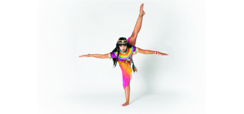 A dancer in classical Egyptian costume stands on one foot with arms outstretched.