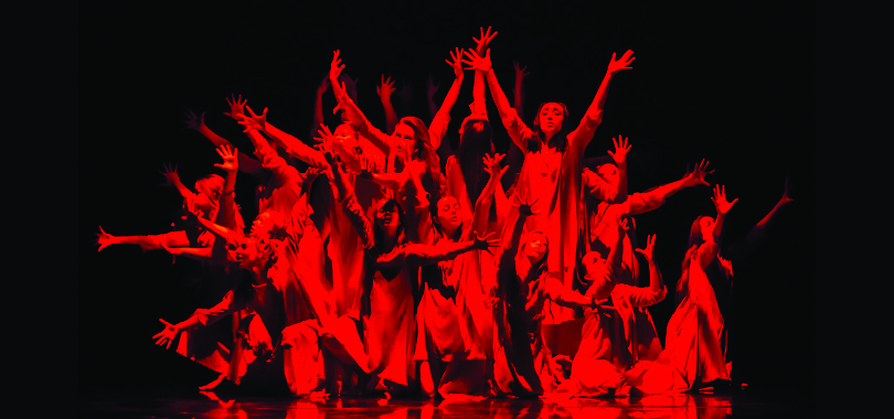 Under red stage lighting, a dozen dancers cluster together with arms reaching up.