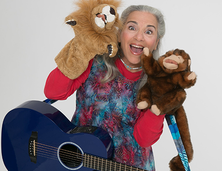 A woman poses with two animal hand puppets and a blue acoustic guitar