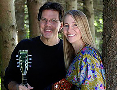 A brown-haired man and a blonde woman pose with an acoustic guitar and a concertina.