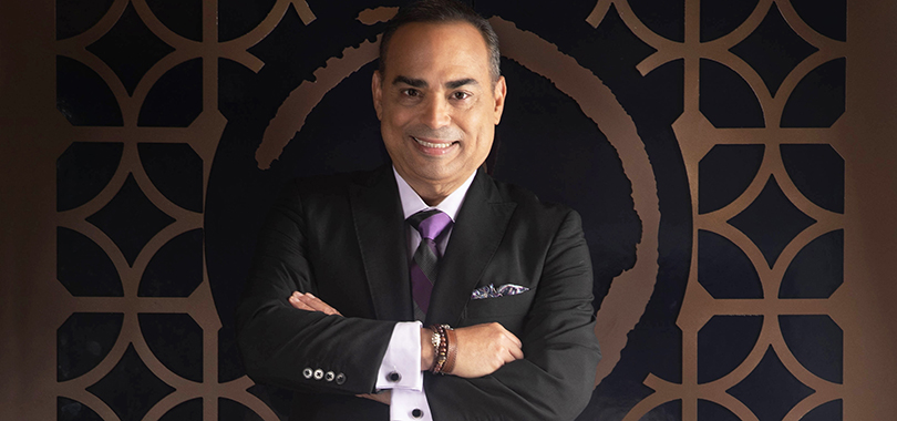 Gilberto Santa Rosa, a Puerto Rican man in his 50s, poses in a black suit with arms crossed, displaying black cufflinks and a leather wrist cuff.