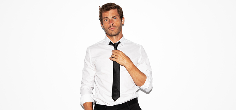 Jon McLaughlin, a white man in his 30s, poses casually against a white wall wearing shirt and tie with his sleeves rolled to the elbows.