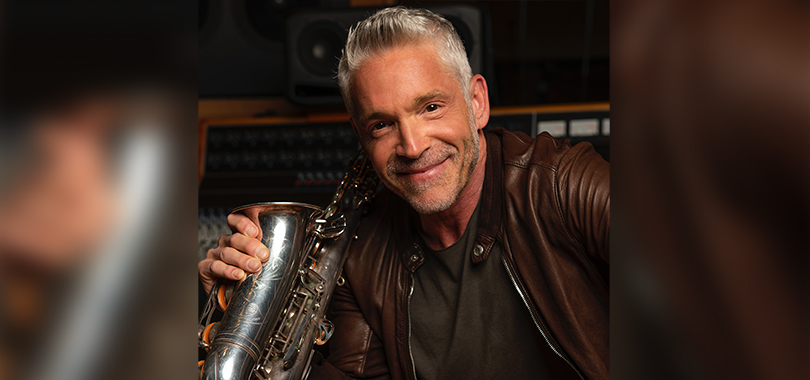 Dave Koz, a white man in his 50s with short gray hair and a mustache, wears a leather jacket over a black t-shirt and poses with his saxophone.