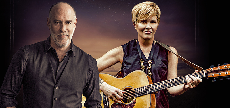Marc Cohn and Shawn Colvin, a white man and woman in their 60s, pose side by side against a backdrop painted like a sky at dusk. Ms. Colvin holds an acoustic guitar.