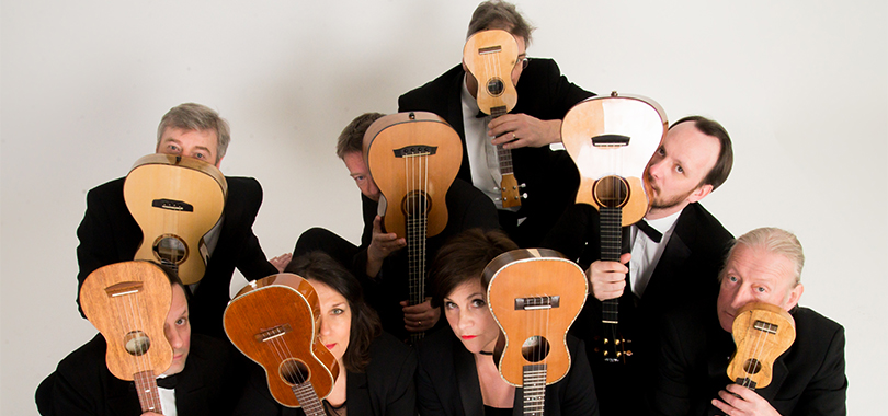 Eight musicians hold their ukuleles up in front of their faces, some fully hidden, some peaking out slightly.