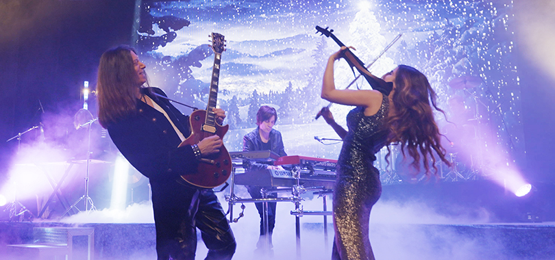 Luminare Christmas An electric guitarist, keyboard player and violinist perform under purple stage lighting against a snowy forest projected background.