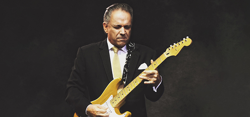 Jimmie Vaughan, a white man in his 60s, wears a black suit with a gold tie. He plays his Fender Stratocaster guitar with eyes closed.