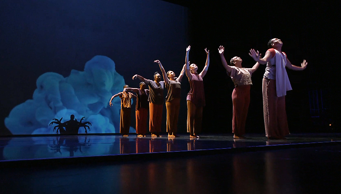 Seven costumed dancers form a line in front of a blue video projection of billowing smoke.