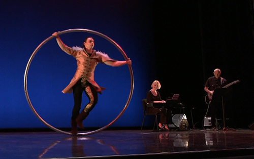 A costumed young man performs on a Cyr wheel, backed by a woman on piano and a man on guitar.