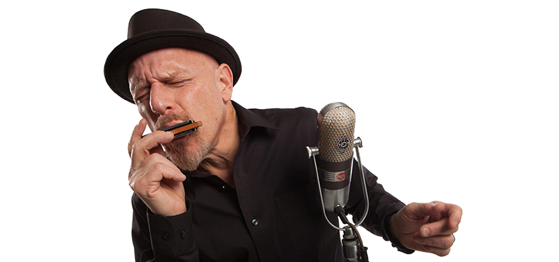 Tad Robinson plays harmonica in front of a large microphone and wears a black shirt with black short-brim hat.