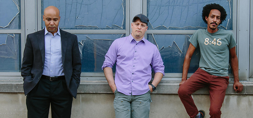 Three men pose casually in front of a building with shattered glass windows.