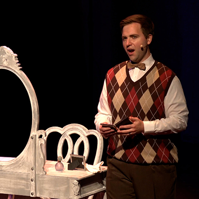 An actor in an argyle sweater sings onstage
