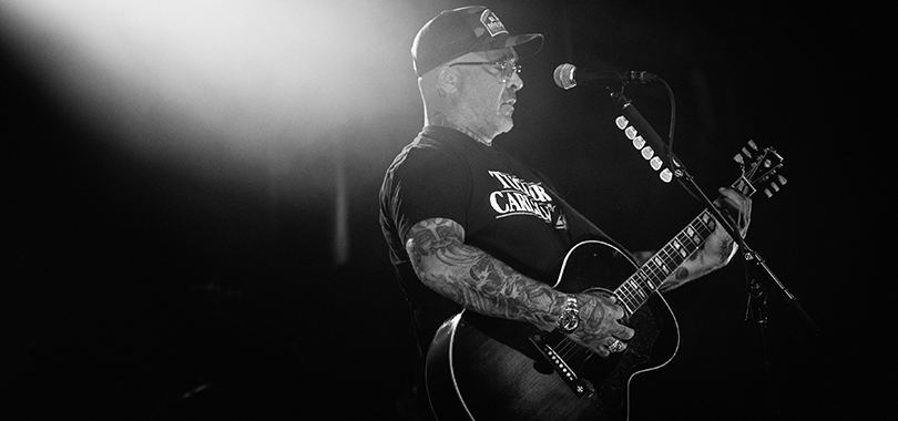 Aaron Lewis, a white man in his 50s, plays acoustic guitar and wears a black t-shirt and ballcap. His arms display multiple tattoos.