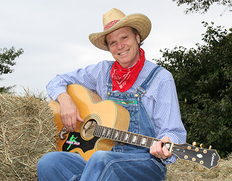 A man dressed as a farmer with denim overalls, a red bandana, and a straw hat plays acoustic guitar next to a hay bale.