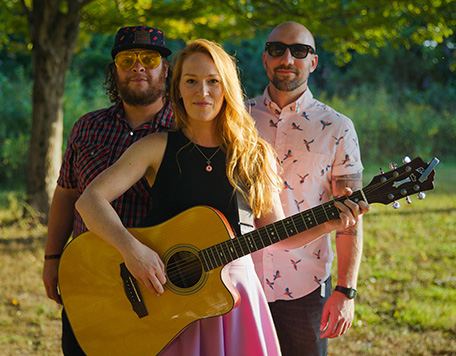 Two men and a woman in trendy clothes and sunglasses pose in a sunny field. The woman holds an acoustic guitar.