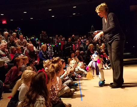 A woman dressed in black performs with a marionette puppet in front of an audience of kids and parents.