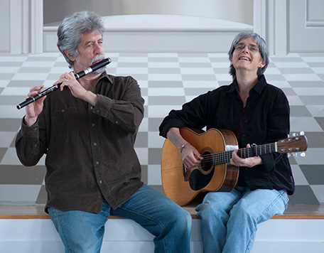 Cindy Kallet plays acoustic guitar and sings while Grey Larsen plays a flute on the edge of a checkered stage.