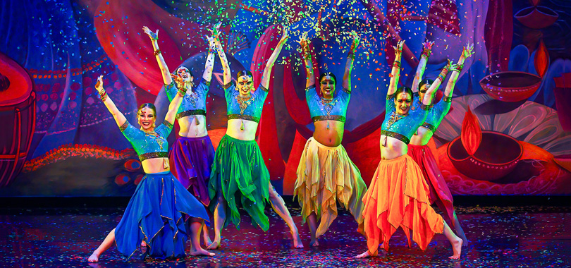 Six women dancers in colorful costumes throw confetti into the air in front of a painted backdrop.