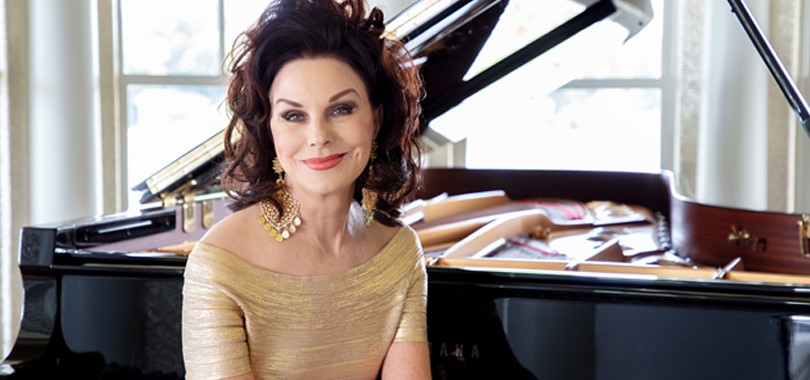Lorie Line wears a gold dress and poses seated in front of a grand piano.