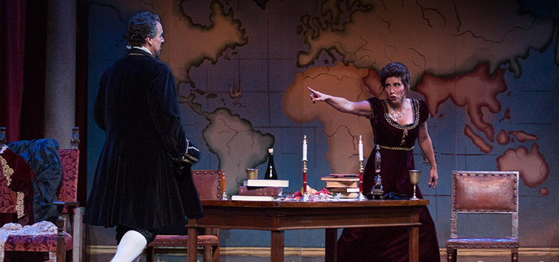A performance photo of Tosca showing two performers in French revolution era costuming. A woman points dramatically at a man across a table against a backdrop of a world map.