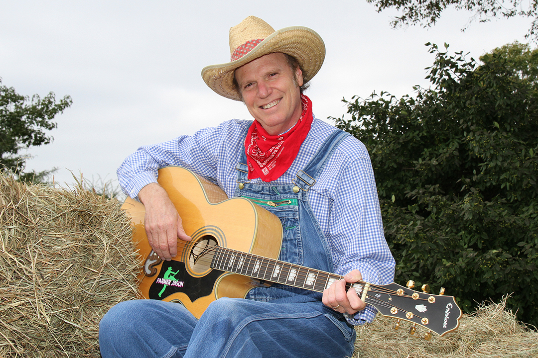 A smiling man in a straw cowboy hat, blue shirt and denim overalls sits on a haystack playing an acoustic guitar.