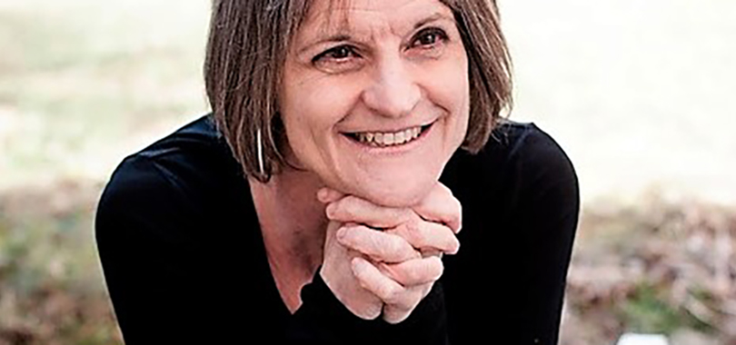 Luminaries: Feathered Tales Sally Perkins has shoulder-length brown hair wears a black top. She smiles and poses with her hands clasped under her chin.