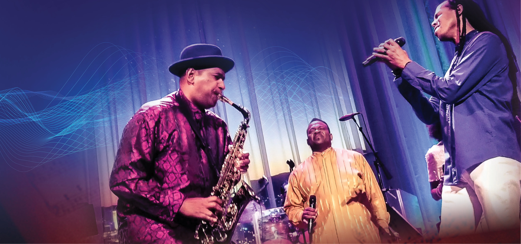 Three men wear colorful clothing and perform on stage, one playing saxophone, two singing into handheld microphones.