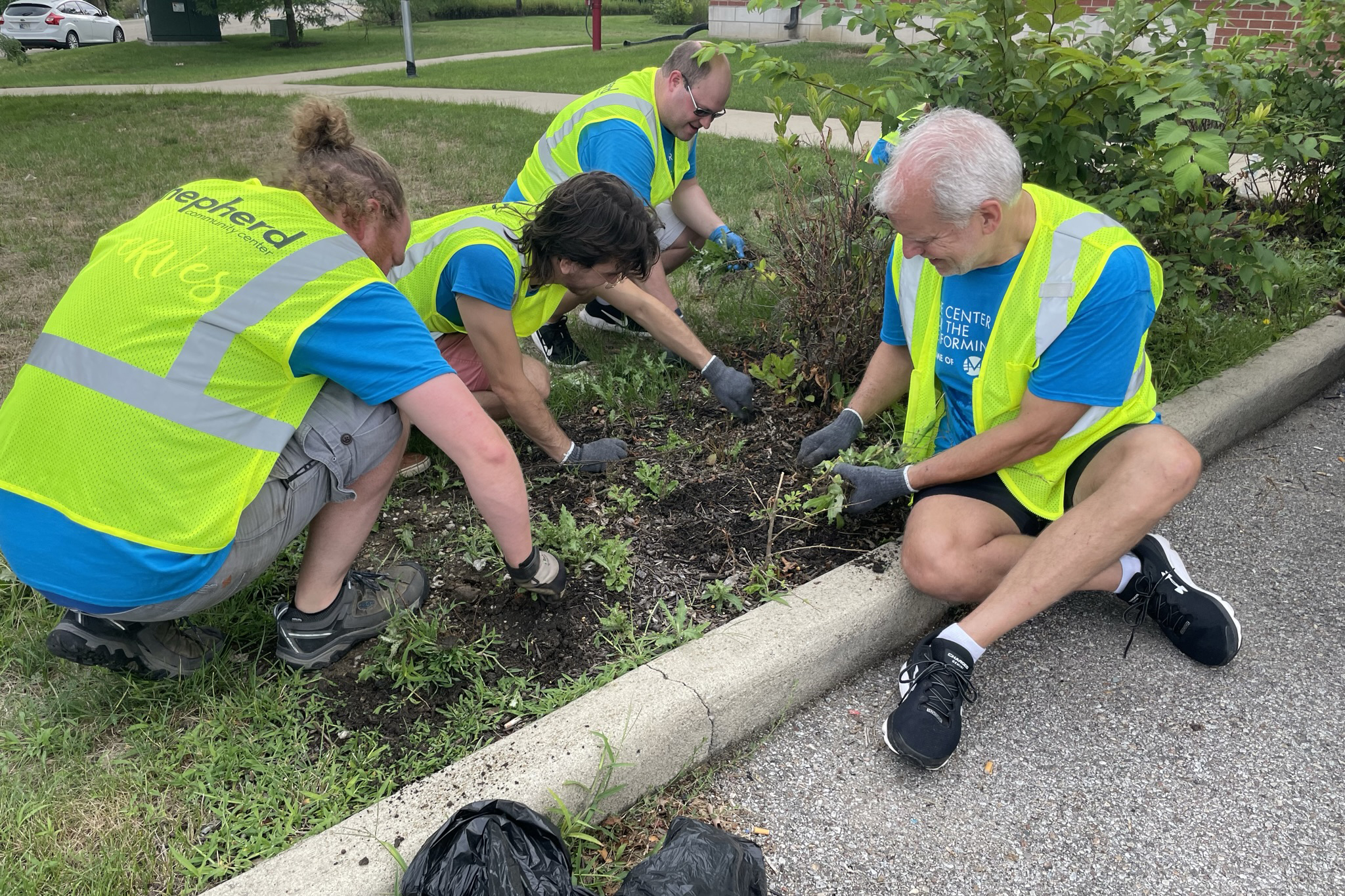 Four men wearing bright vests pull weeds near a parking lot.